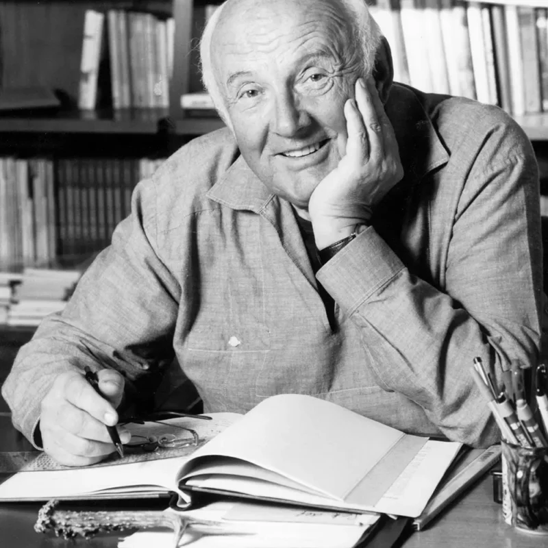 Otfried Preußler sitzt an einem Tisch vor einem aufgeschlagenen Notizbuch, auf dem seine Brille liegt. Er hält einen Stift in der Hand und lächelt in die Kamera. Im Hintergrund ist verschwommen eine Bibliothek zu erkennen.