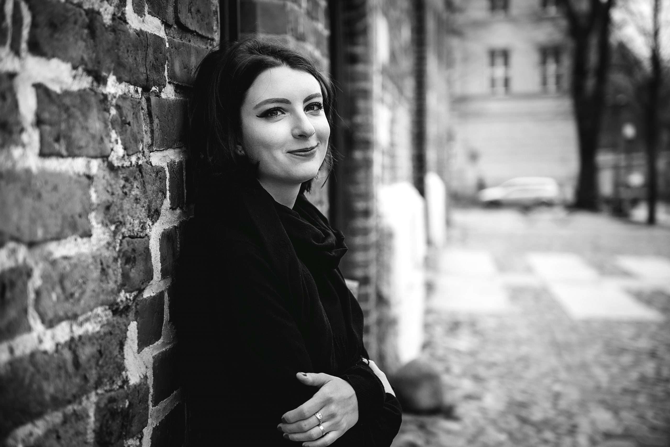 Schwarz-weiß Fotografie einer jungen Frau in Schwarz, im Profil an einer Mauer lehnend mit verschränkten Armen.