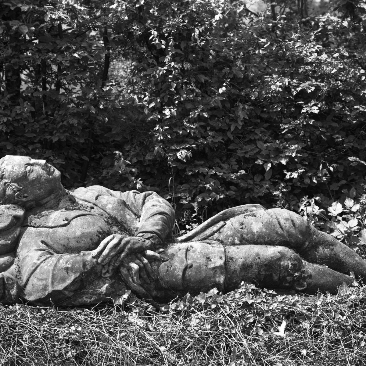 Ein Schwarz-Weiß-Foto einer liegenden Statue, die eine Figur in altertümlicher Kleidung mit gekreuzten Armen darstellt, eingebettet in Gras und umgeben von Bäumen.
