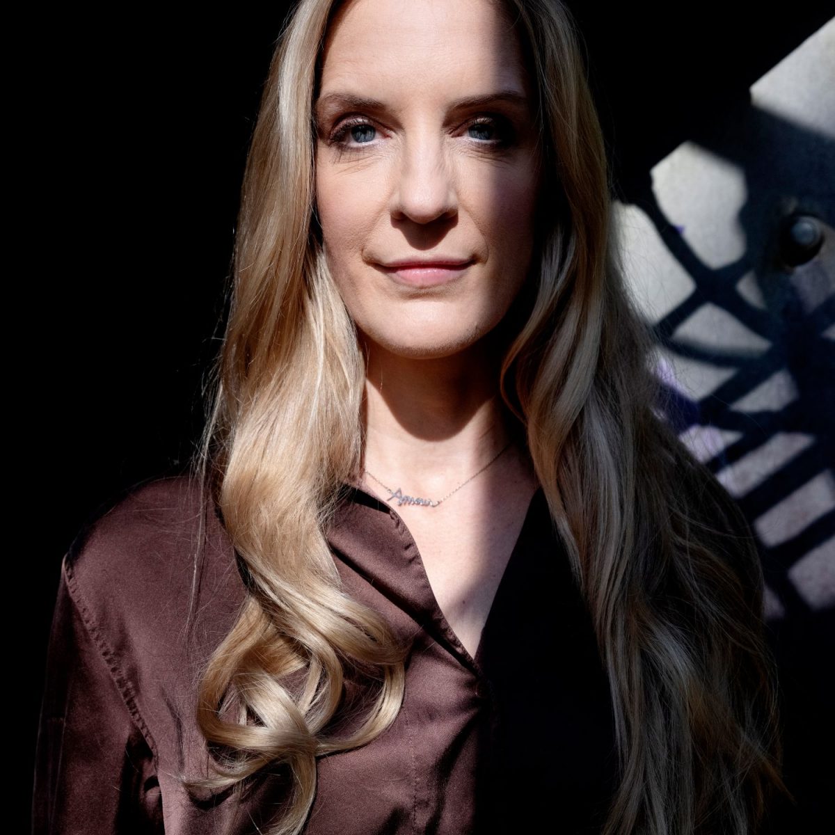 Ein Porträt von Caro Matzko, einer blonden Frau mit blauen Augen, die eine braune Satinbluse und eine Halskette trägt. Ihr Gesicht wird teilweise von einem Schattenmuster bedeckt.