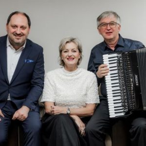 Drei Künstler auf einem Sofa sitzend. Herr Zdeněk Hazdra, Frau Sylva Čmugrová und Herr Ladislav Horák mit einem Akkordeon.