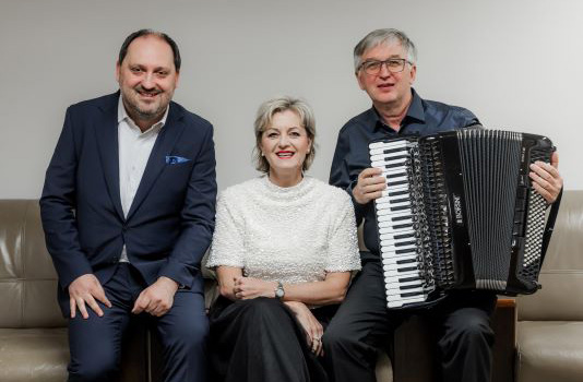 Drei Künstler auf einem Sofa sitzend. Herr Zdeněk Hazdra, Frau Sylva Čmugrová und Herr Ladislav Horák mit einem Akkordeon.