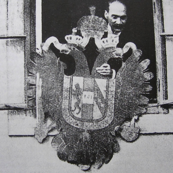 Schwarzweißfotografie eines Mannes im geöffneten Fenster, der das österreichische Wappen mit dem Doppeladler in der Hand hält, im Begriff, es aus dem Fenster fallen zu lassen.