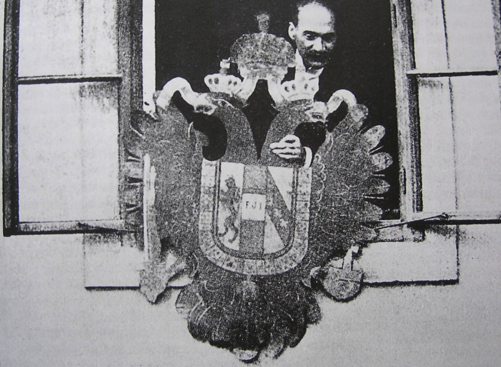 Schwarzweißfotografie eines Mannes im geöffneten Fenster, der das österreichische Wappen mit dem Doppeladler in der Hand hält, im Begriff, es aus dem Fenster fallen zu lassen.