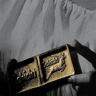 Schwarzweiß-Foto mit gold eingefärbtem Stück Seife, links und rechts jeweils zwischen zwei Fingern gehalten. Auf der Seife ist ein Hirsch und der Schriftzug 