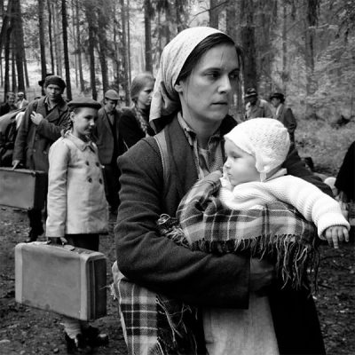 Schwarzweiß-Foto, auf dem eine Frau ein kleines Kind auf dem Arm hält. Im Hintergrund sind weitere Kinder und Erwachsene mit Gepäck in den Händen