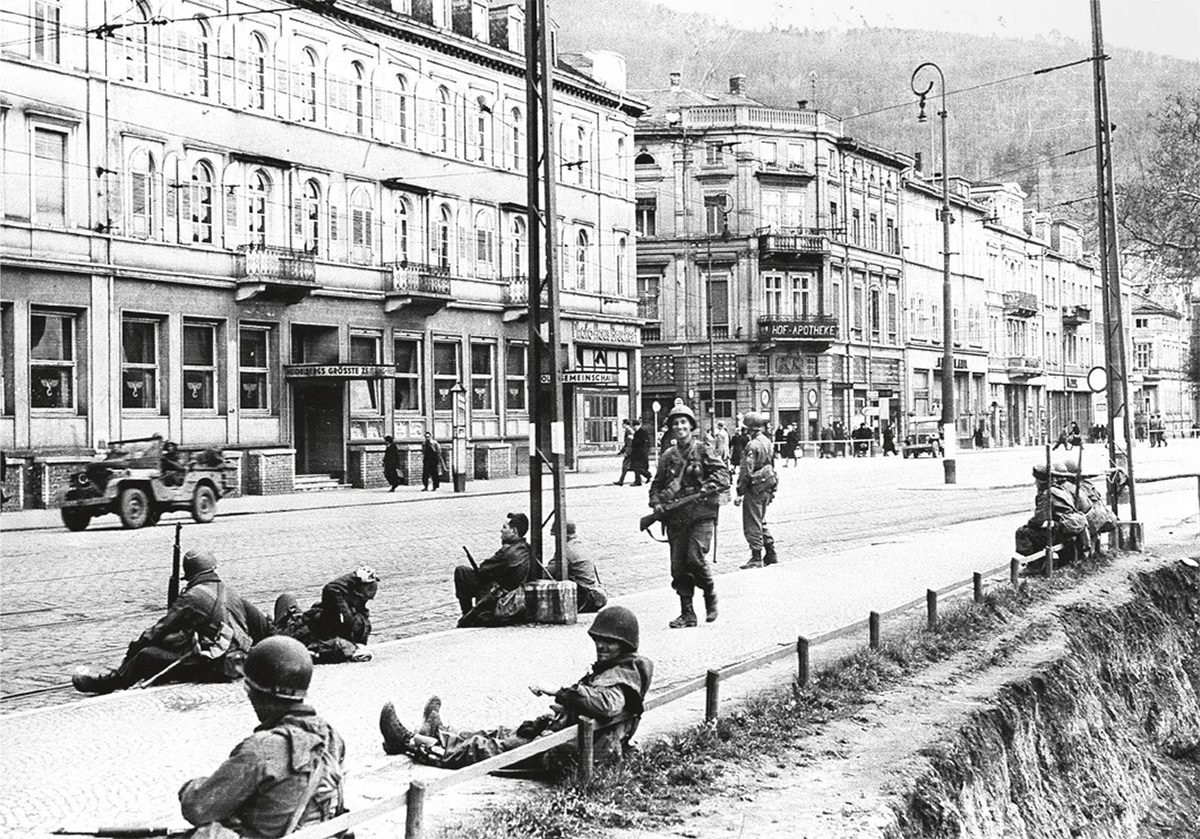 Weinheim_Foto_Archiv Miloš Doležal Schwarzweiß-Foto einer Stadt mit Hausfassaden und einer Straße, auf der sich Soldaten befinden, überwiegend auf der Straße sitzend